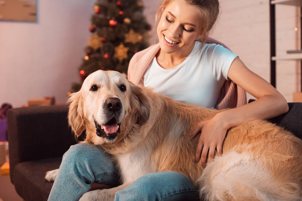 beautiful smiling young blonde woman sitting on couch and hugging golden retriever dog at christmas time - Фото, изображение