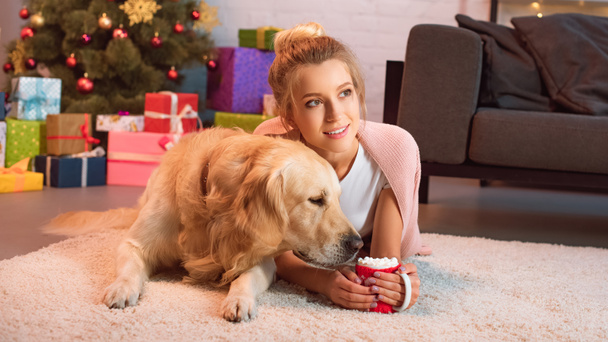 belle jeune femme blonde couchée sur le sol avec chien récupérateur d'or et tasse de cacao chaud avec guimauves à Noël
  - Photo, image