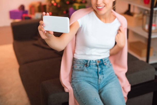 обрезанный вид улыбающейся молодой женщины, сидящей на диване и делающей селфи на смартфоне на Рождество
  - Фото, изображение