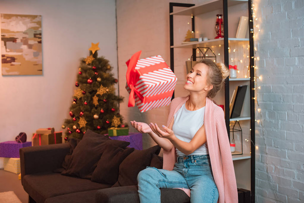 cheerful young blonde woman sitting on couch and throwing up present in air at christmas time  - Foto, Imagen