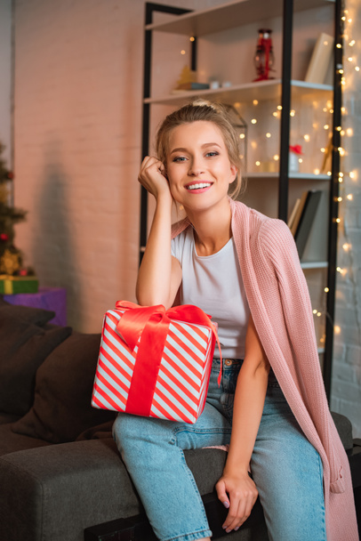 happy young blonde woman sitting on couch with present and looking at camera at christmas time  - Фото, изображение