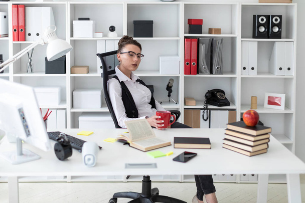 Młoda dziewczyna siedzi przy biurku komputer w biurze i w jej ręce trzyma kubek czerwony. - Zdjęcie, obraz