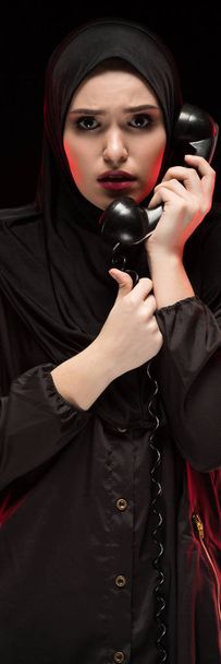 Porträt einer schönen, verängstigten jungen Muslimin, die einen schwarzen Hijab trägt und auf schwarzem Hintergrund um Hilfe ruft - Foto, Bild