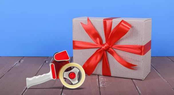 Portó és csomagolás házhozszállítás - kis csomag megkötött megjelöl egy piros orr és a szalag adagoló a egy barna fa és a kék háttér. - Fotó, kép