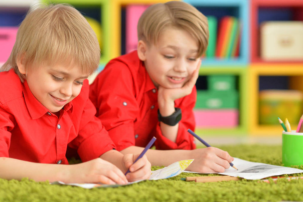 Два улыбающихся мальчика в красных рубашках рисуют карандашами на полу с зеленым ковром
 - Фото, изображение
