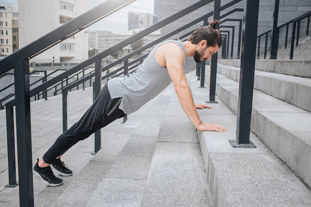 Picture of well-built athlete exercising on steps. He stands in plank position and looks down. Guy is calm and concentrated. He is alone there. - Foto, Imagen