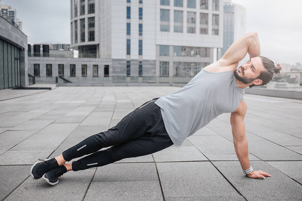 Picture of well-bilt young man stretching his body by doing exercises. He holds one hand behind head. Guy looks forward. He is concentated. Young man exercises alone. - Foto, Bild