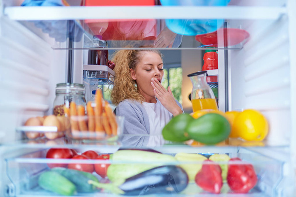 Young Caucasian woman yawning and taking juice from fridge. Morning routine. Picture taken from the inside of fridge. - Photo, Image