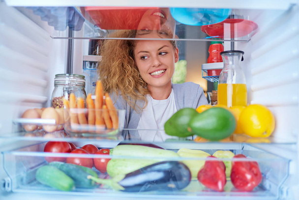 Жінка стоїть перед холодильником, повним продуктових магазинів і шукає щось попоїсти. Картина, зроблена зсередини холодильника
. - Фото, зображення