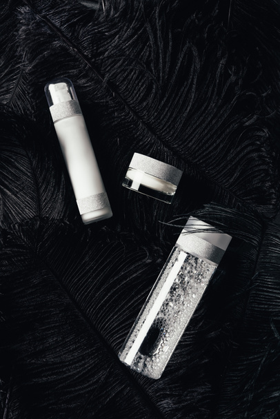 bovenaanzicht van schoonheid crème, micellaire water en lotion op oppervlak met zwarte veren  - Foto, afbeelding