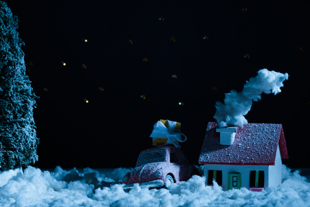 Nahaufnahme von Spielzeugauto mit Weihnachtsgeschenk und schneebedecktem Haus in der Nacht - Foto, Bild