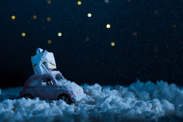 Nahaufnahme eines Spielzeugautos mit Geschenkbox, das in der Nacht im Schnee steht, Weihnachtskonzept - Foto, Bild