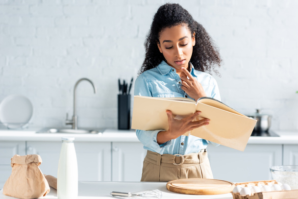 pensive attractive african american girl looking at cookbook in kitchen - Fotoğraf, Görsel