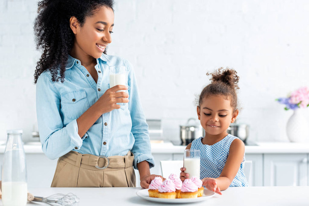 african american mother and daughter standing with glasses of milk and cupcakes in kitchen - Φωτογραφία, εικόνα