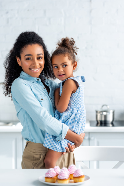 happy african american mother hugging daughter in kitchen, cupcakes on table - Photo, Image