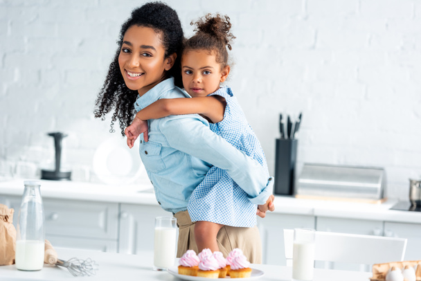 afro-américaine mère donnant piggyback à fille dans la cuisine, cupcakes maison sur la table
 - Photo, image