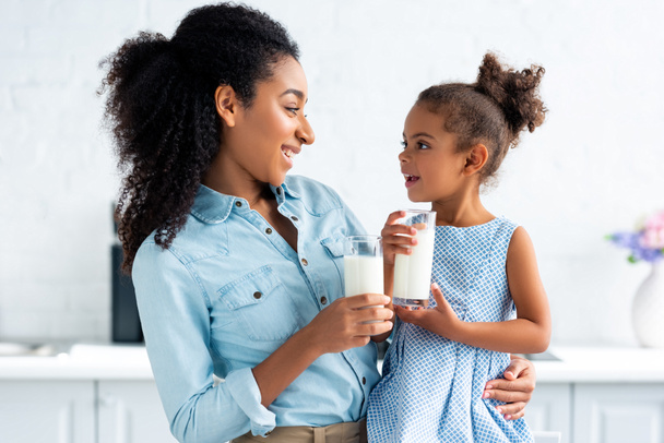 χαρούμενα αφρικανική αμερικανική μητέρα και κόρη κρατώντας ποτήρια γάλα στην κουζίνα και να βλέπουν ο ένας τον άλλον - Φωτογραφία, εικόνα
