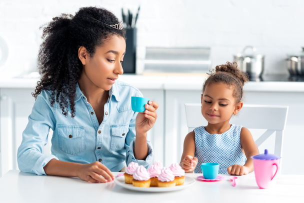 african american mother and daughter with tiaras sitting at table with cupcakes in kitchen - 写真・画像