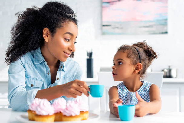 african american mother and daughter with tiaras holding plastic cups and looking at each other in kitchen - Foto, Bild