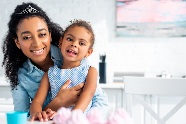 smiling african american mother hugging daughter at table with cupcakes in kitchen - 写真・画像