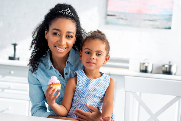 afro-americana mãe abraçando filha com cupcake na cozinha, olhando para a câmera
 - Foto, Imagem