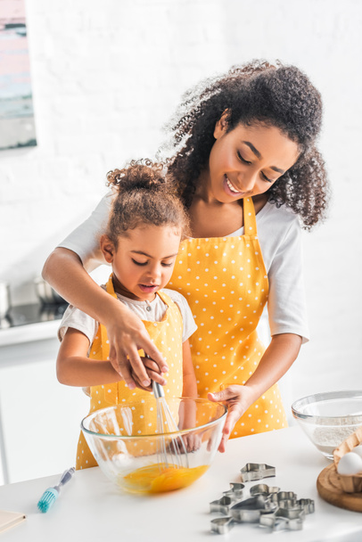 african american mother and daughter whisking eggs for dough together in kitchen - Photo, Image