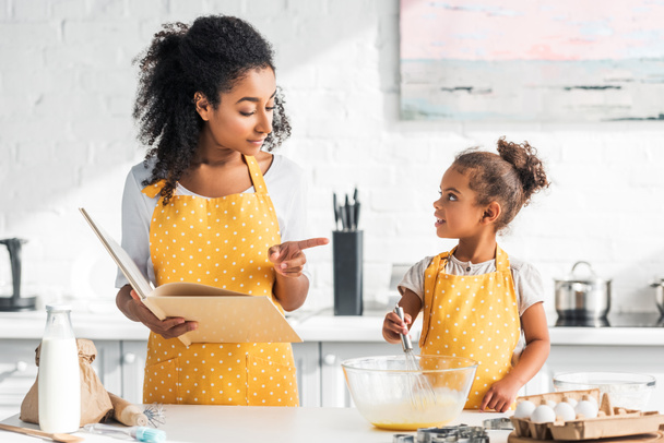 Afrikkalainen amerikkalainen äiti tilalla keittokirja ja osoittaa jotain tytär valmistelee taikina keittiössä
 - Valokuva, kuva