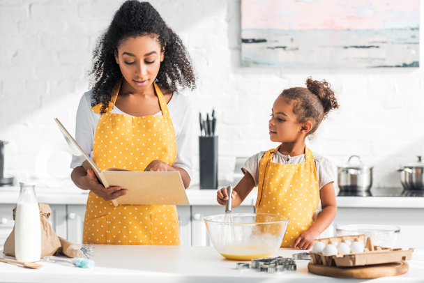 madre afroamericana leyendo libro de cocina e hija preparando masa en la cocina
 - Foto, imagen