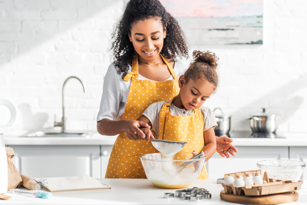 smiling african american mother and daughter preparing dough and sieving flour in kitchen - Photo, Image