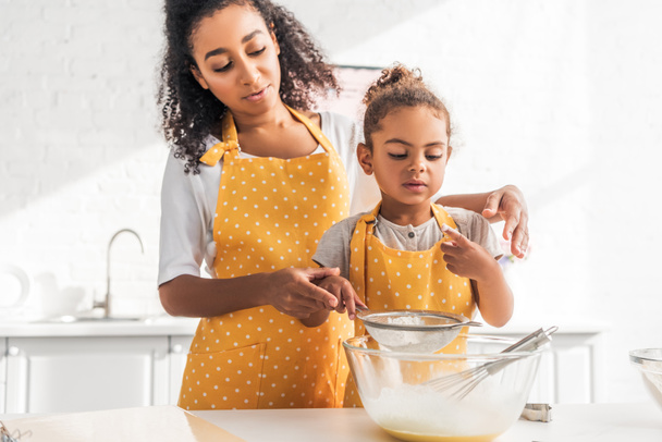 madre e hija afroamericanas preparando masa y tamizando harina juntas en la cocina
 - Foto, Imagen