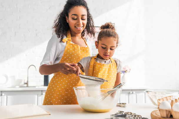 Африканская американская мать помогает дочери готовить тесто и просеивать муку на кухне
 - Фото, изображение