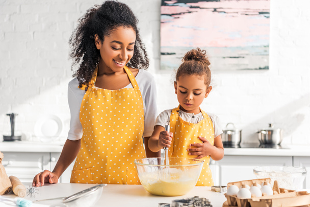 Африканская американская дочь готовит и взбивает тесто на кухне, улыбаясь матери глядя на миску
 - Фото, изображение