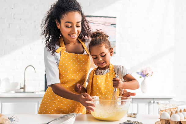 hymyilevä afrikkalainen amerikkalainen äiti auttaa tytärtä valmistelemaan ja vatkaamaan taikinaa keittiössä
 - Valokuva, kuva