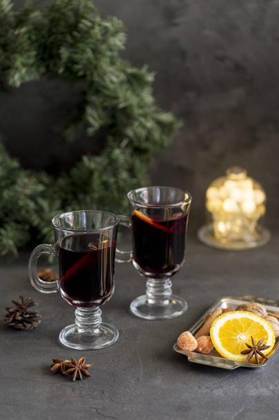 Winter composition. Mulled wine in glasses at black background. Fir wreath, lights, tray with orange, cinnamon, nuts, cone and spices near. - Φωτογραφία, εικόνα
