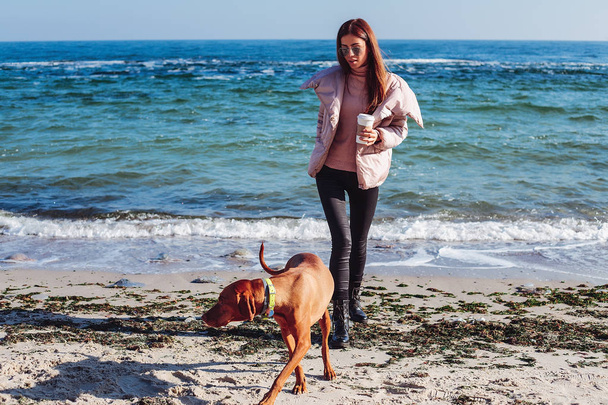 Young woman playing with dog and holding cup of coffee on seacoast - Photo, Image