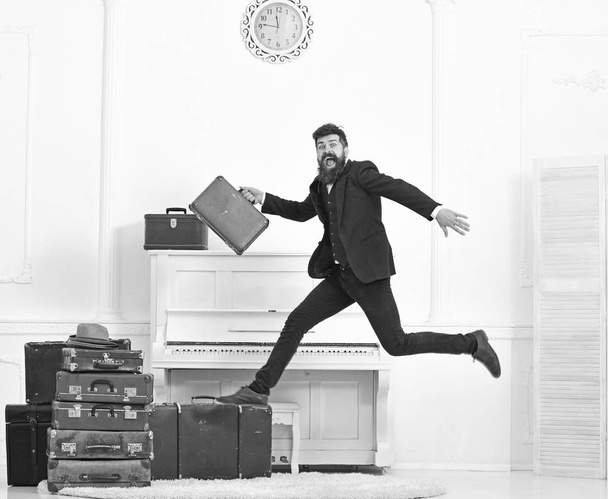 Macho attractive, elegant on cheerful face carries vintage suitcases, jumping. Man with beard and mustache in suit carries luggage, luxury white interior background. Long awaited vacation concept - Zdjęcie, obraz