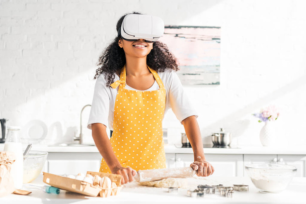chica afroamericana en delantal y realidad virtual auriculares balanceo masa con rodillo en la cocina
 - Foto, Imagen