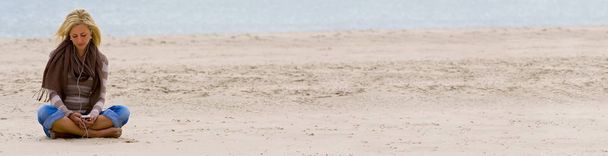 Web banner panoramic photograph of beautiful sad depressed young blond woman or girl sitting alone on a beach listening to music on headphones and smart phone - Fotó, kép