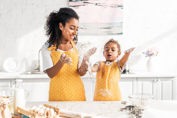 african american mother and daughter in aprons preparing dough and having fun with flour in kitchen - Foto, immagini