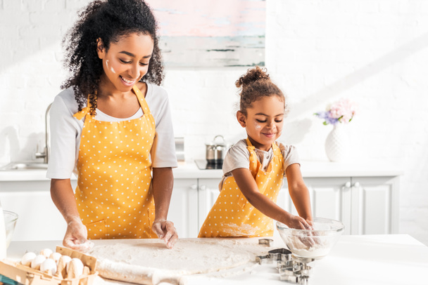 glimlachend Afro-Amerikaanse moeder en dochter voorbereiding deeg samen in de keuken - Foto, afbeelding