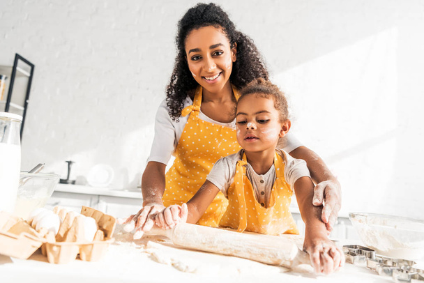 feliz afro-americana mãe ajudando filha rolando massa com rolo de pino na cozinha
 - Foto, Imagem