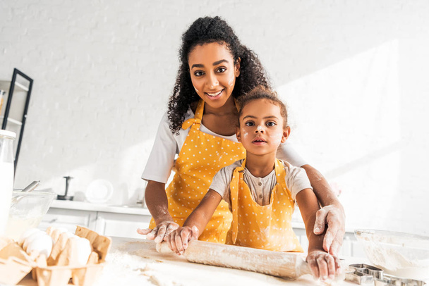 afro-americana mãe ajudando filha rolando massa com rolo de pino na cozinha e olhando para a câmera
 - Foto, Imagem