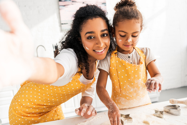 camera point of view of smiling african american mother and daughter preparing cookies with molds in kitchen - Foto, Imagen