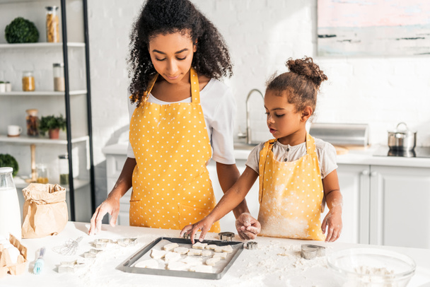 african american mother and daughter in yellow aprons looking at cookies on tray in kitchen - Photo, Image