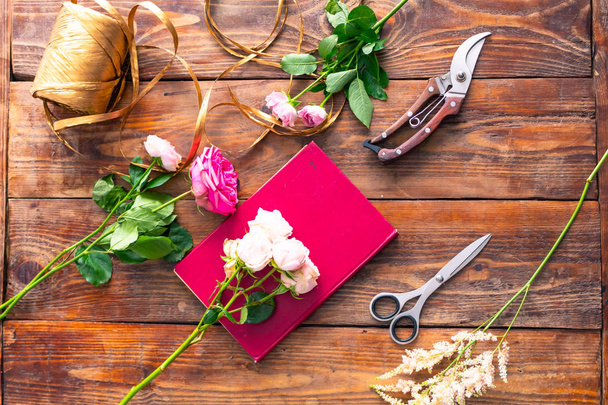 Στην ξύλινη επιφάνεια ορίζονται εργαλεία για ανθοδέσμες λουλουδιών: τριαντάφυλλα, χρωματιστά κορδέλα, ψαλίδι, τριαντάφυλλα. - Φωτογραφία, εικόνα