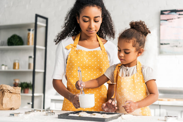 afrikanisch-amerikanische Mutter und Tochter werden Öl auf ungebackene Kekse in der Küche auftragen - Foto, Bild