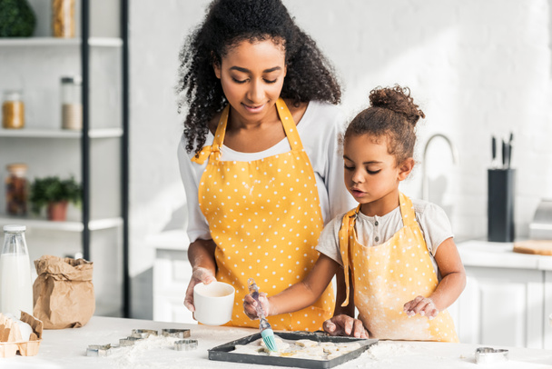 attrayante mère afro-américaine et adorable fille appliquant de l'huile sur les biscuits non cuits dans la cuisine
 - Photo, image
