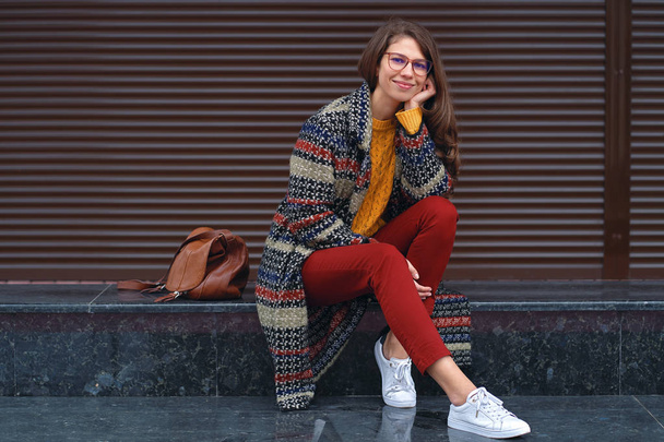 Portrét mladé, krásné ženy, které nosí stylový kabát, červené kalhoty, oranžový svetr, bílé tenisky, s hnědou koženou tašku, pózuje v ulici Evropské město. Kopírovat prostor - Fotografie, Obrázek