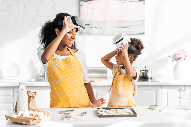 african american mother and daughter preparing dough in virtual reality headsets and looking at each other in kitchen - Valokuva, kuva