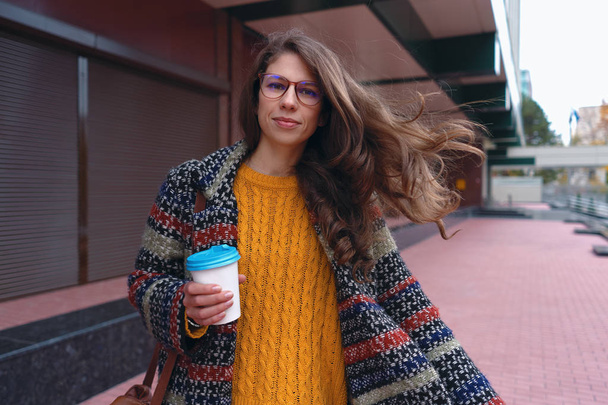 Miejski styl i kawa w podróży. Stylowy i młoda kobieta w płaszcz picia kawy na zewnątrz. Dorywczo dziewczynka w nowoczesny miejski strój i okulary spaceru ulicami miasta. - Zdjęcie, obraz